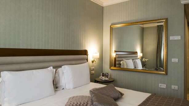 A hotel room with a large bed, a nightstand, a mirror, and a lamp. - Hotel Maggior Consiglio (Treviso)