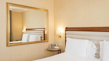 Habitación de hotel con un espejo grande, una cama con sábanas blancas, una mesita de noche pequeña con una lámpara, y una lámpara de pared.
