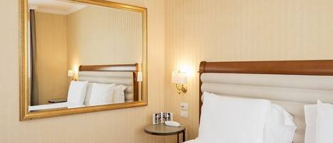 Habitación de hotel con un espejo grande, una cama con sábanas blancas, una mesita de noche pequeña con una lámpara, y una lámpara de pared.