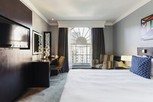 A hotel room with a bed, desk, chair, and a large window with curtains.