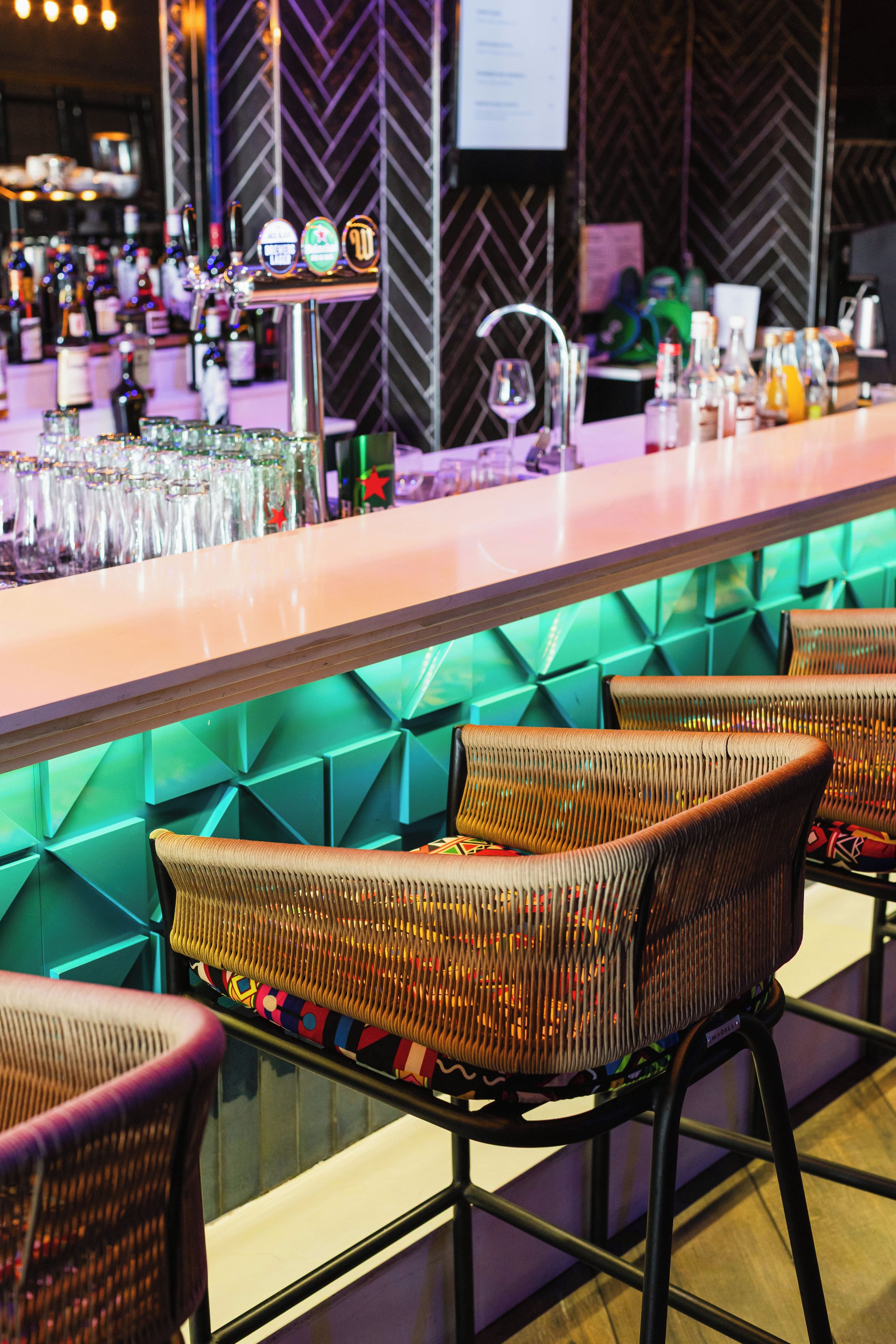Una zona de bar con mostrador, taburetes y una variedad de botellas y vasos.