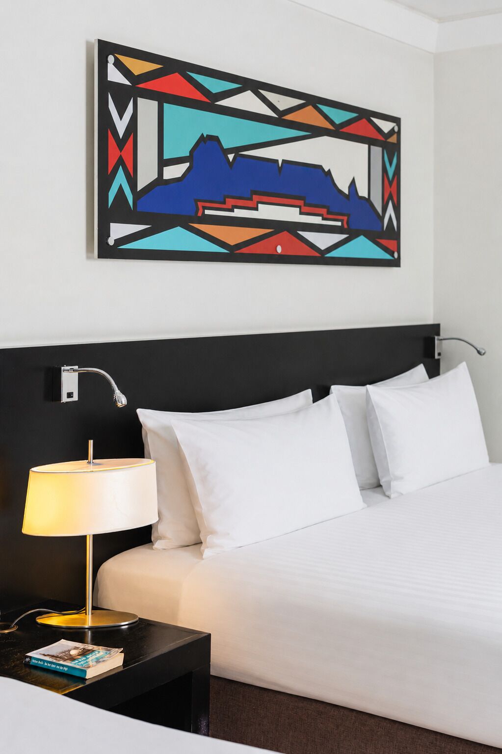 A hotel room with a bed, white pillows, a black nightstand with a lamp, a book, and a colorful abstract painting on the wall.