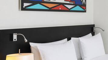 A hotel room with a bed, white pillows, a black nightstand with a lamp, a book, and a colorful abstract painting on the wall.