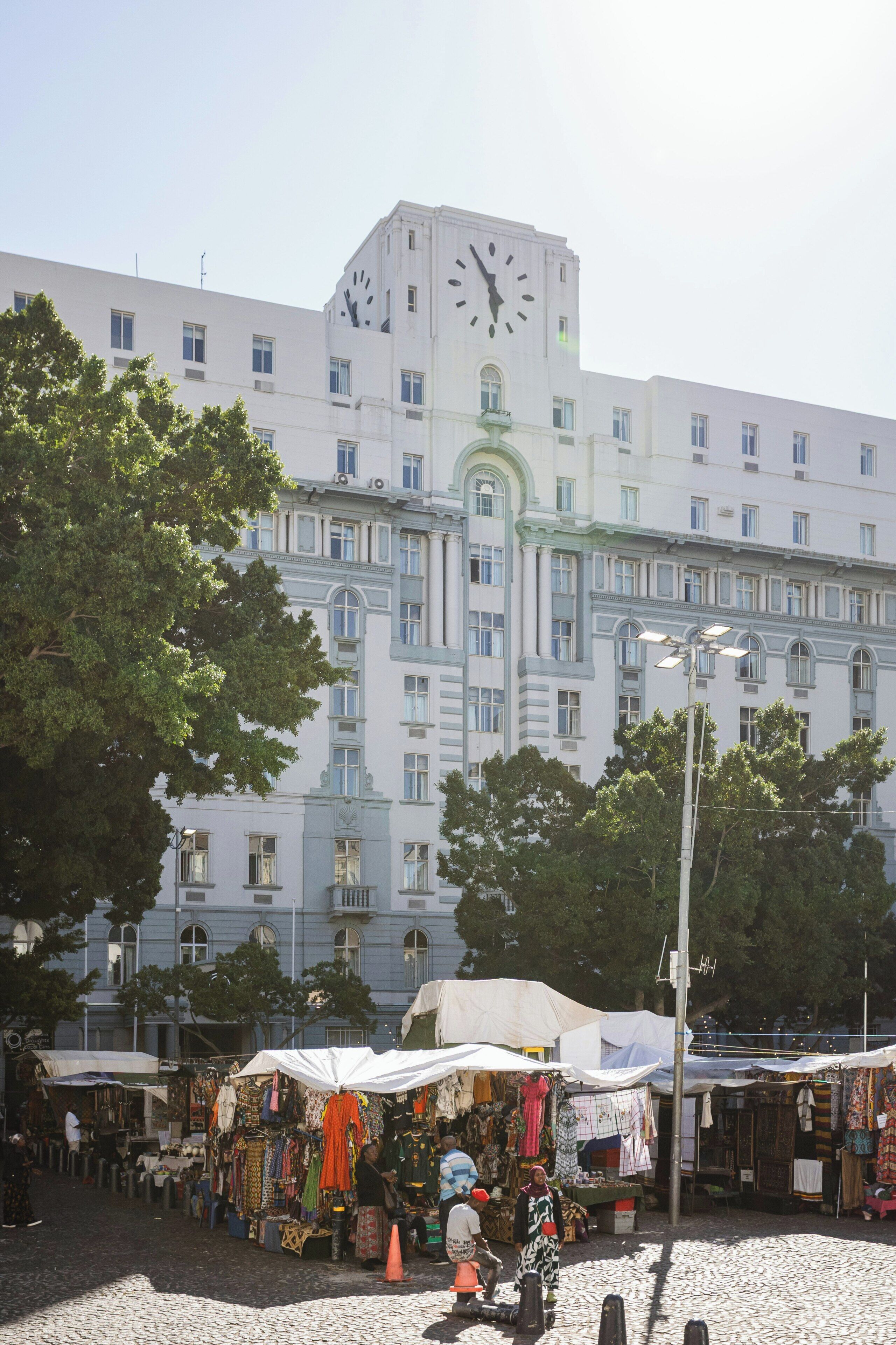 A building with a large clock on the facade, surrounded by trees and a market area with stalls.