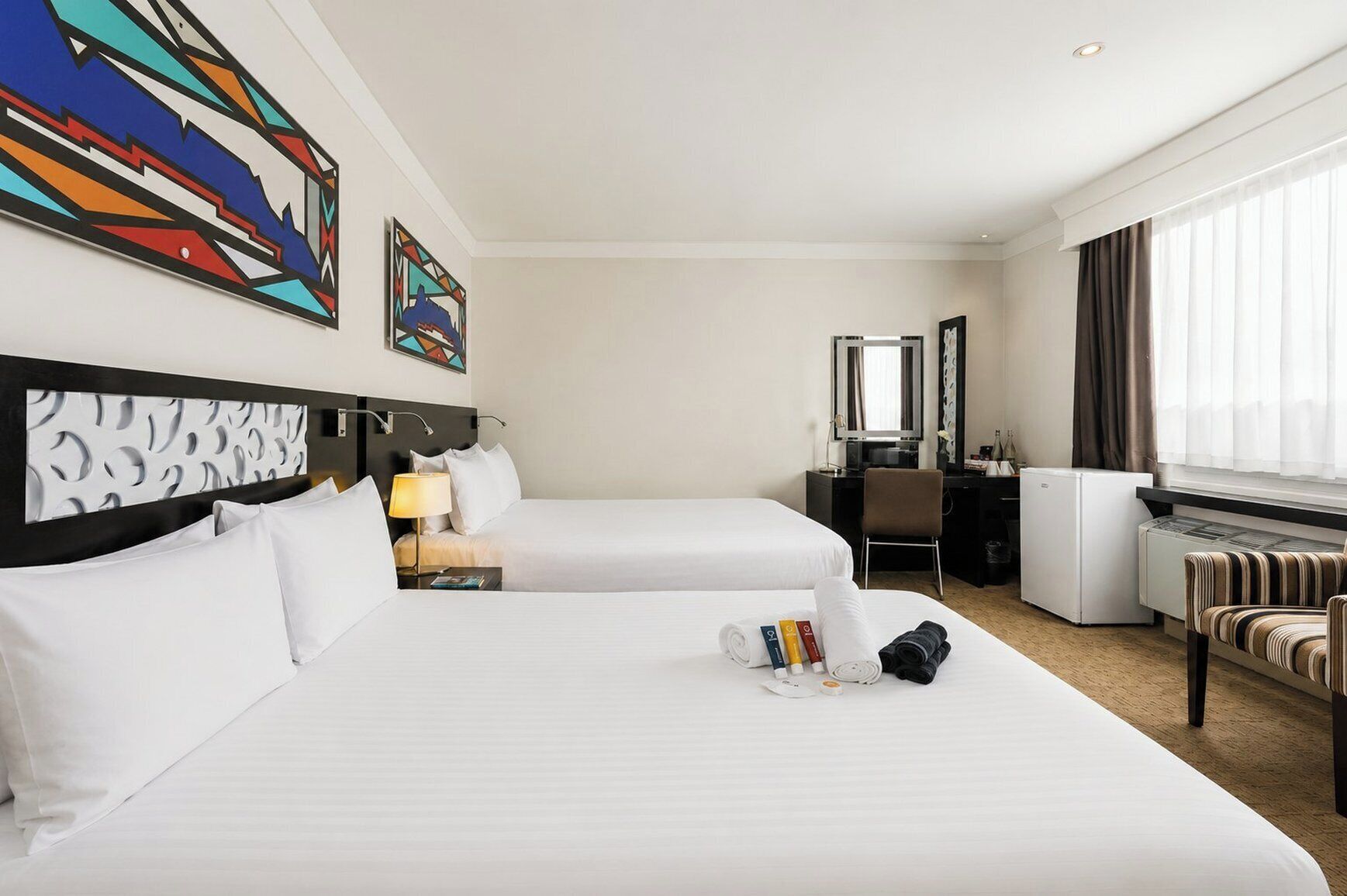 A hotel room with two beds, a desk, a chair, a radiator, and artwork on the walls.