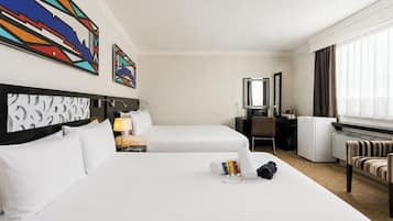 A hotel room with two beds, a desk, a chair, a radiator, and artwork on the walls.