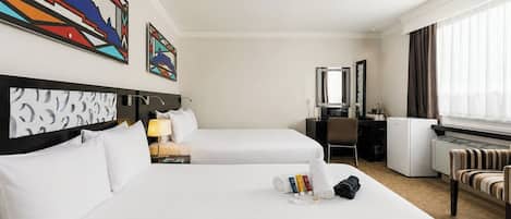 A hotel room with two beds, a desk, a chair, a radiator, and artwork on the walls.
