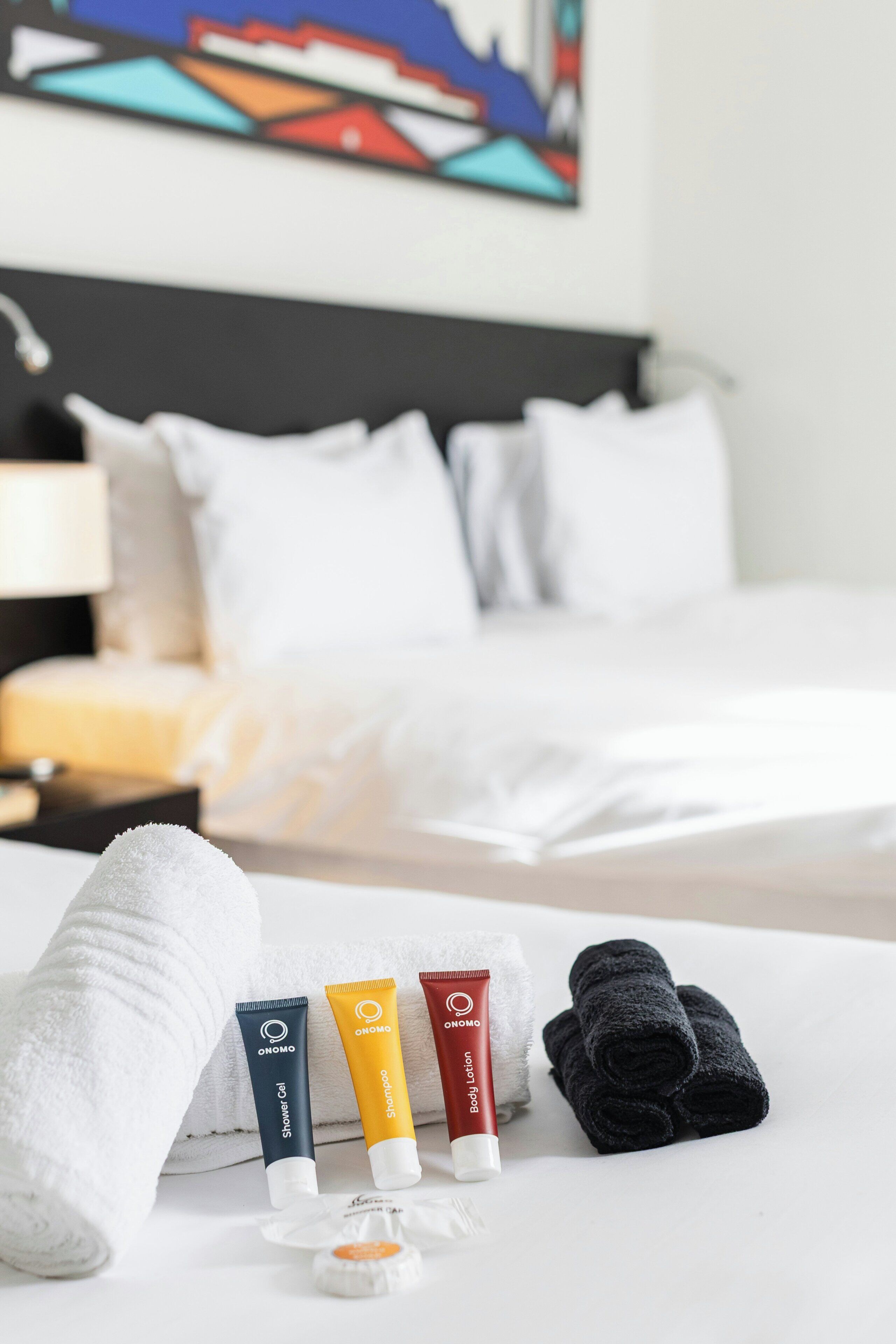 Ein Hotelzimmer mit einem ordentlich bezogenen Bett, einer Zusammenstellung von Pflegeprodukten, darunter Duschgel, Körperlotion und ein Handtuch, sowie einem Bild an der Wand.