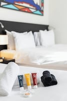 A hotel room with a neatly made bed, a set of toiletries including shower gel, body lotion, and a towel, and a painting on the wall.