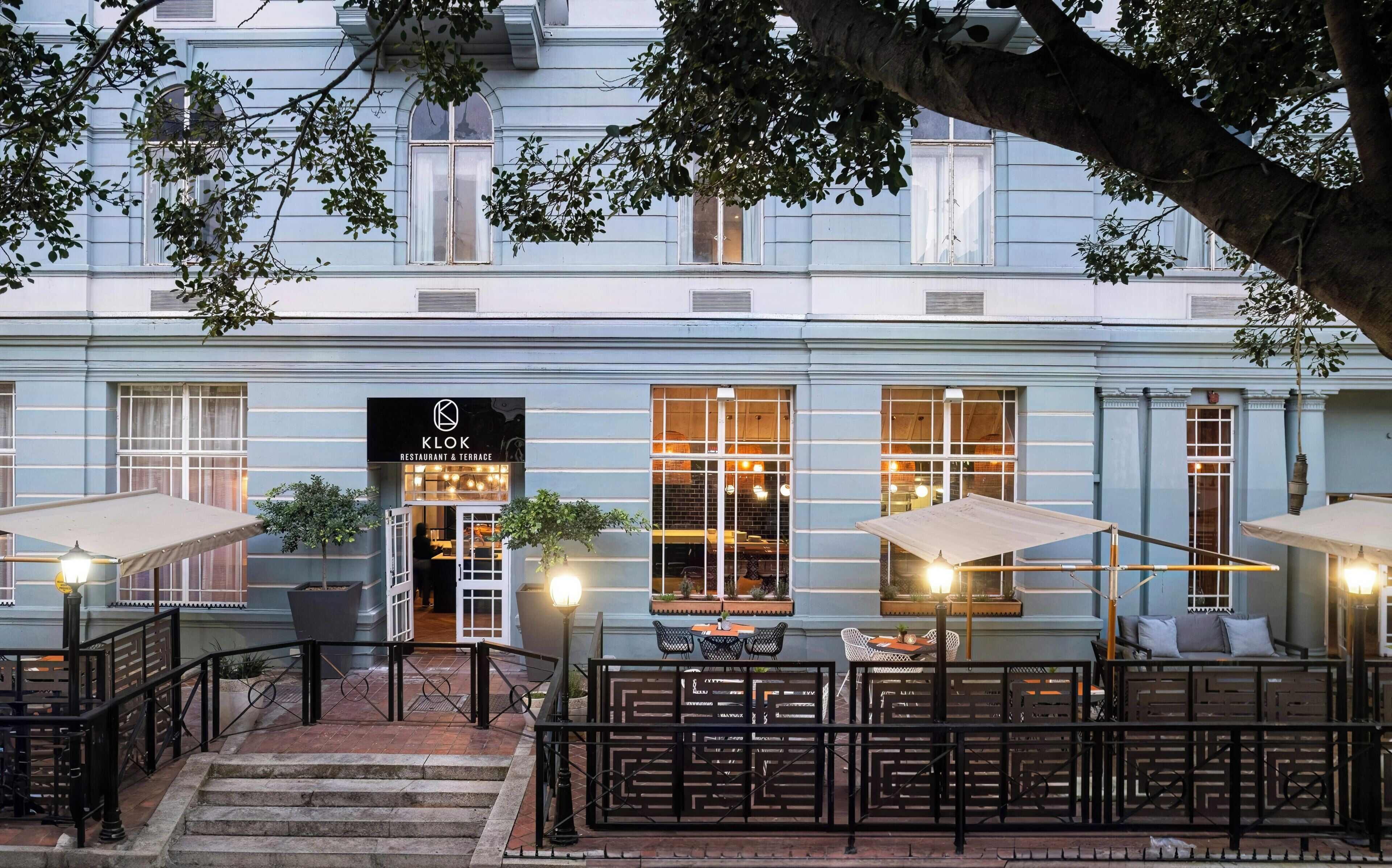 A restaurant terrace with outdoor seating, a staircase leading up to the entrance, and a building facade with large windows.