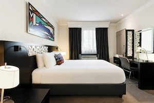 A hotel room with a large bed, a desk, a chair, a desk lamp, a wall-mounted TV, and a window with curtains.