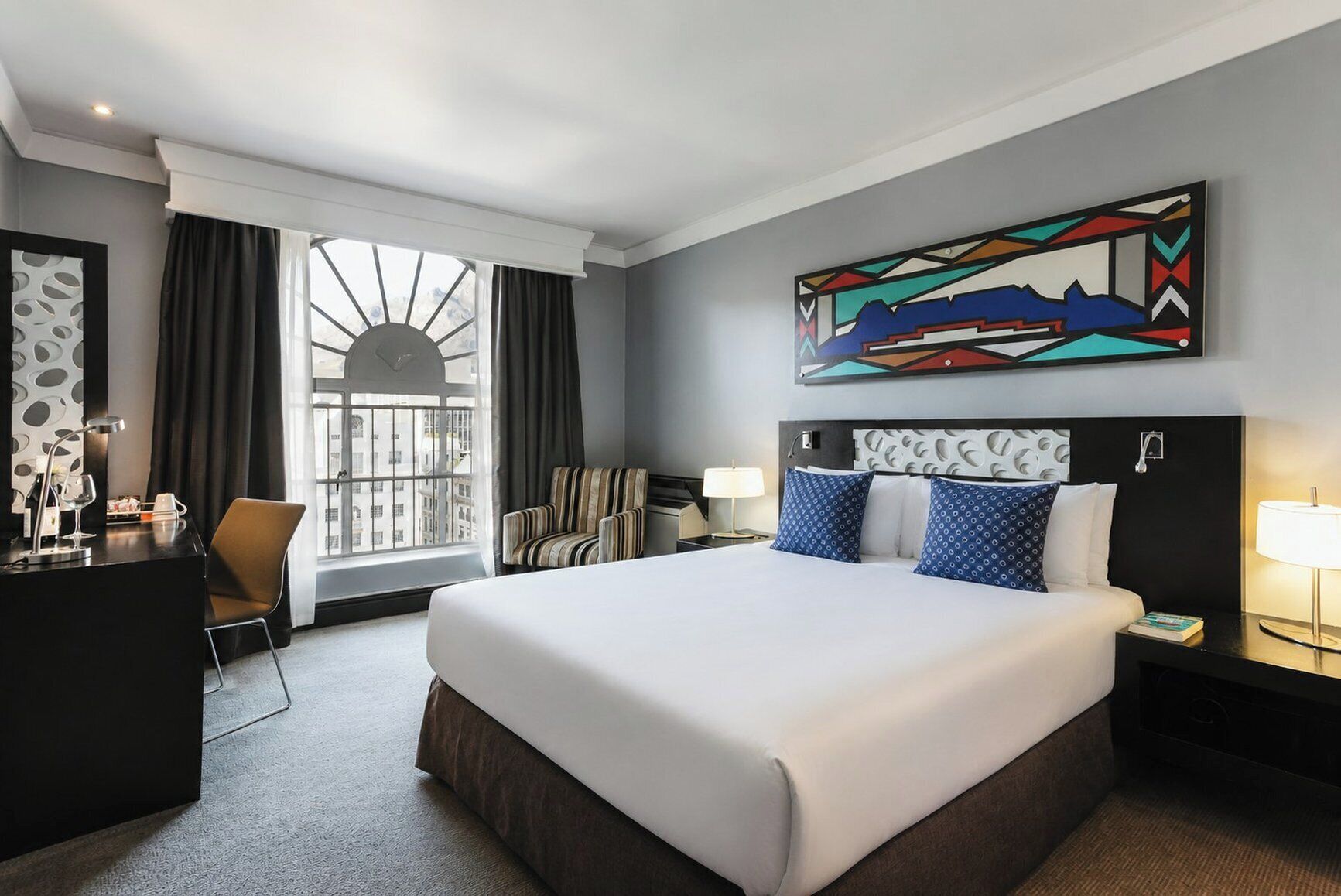 A hotel room with a large bed, a desk with a chair, a window with curtains, and a painting on the wall.
