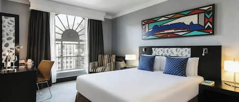 Une chambre d’hôtel avec un grand lit, un bureau avec une chaise, une fenêtre avec des rideaux et un tableau au mur.