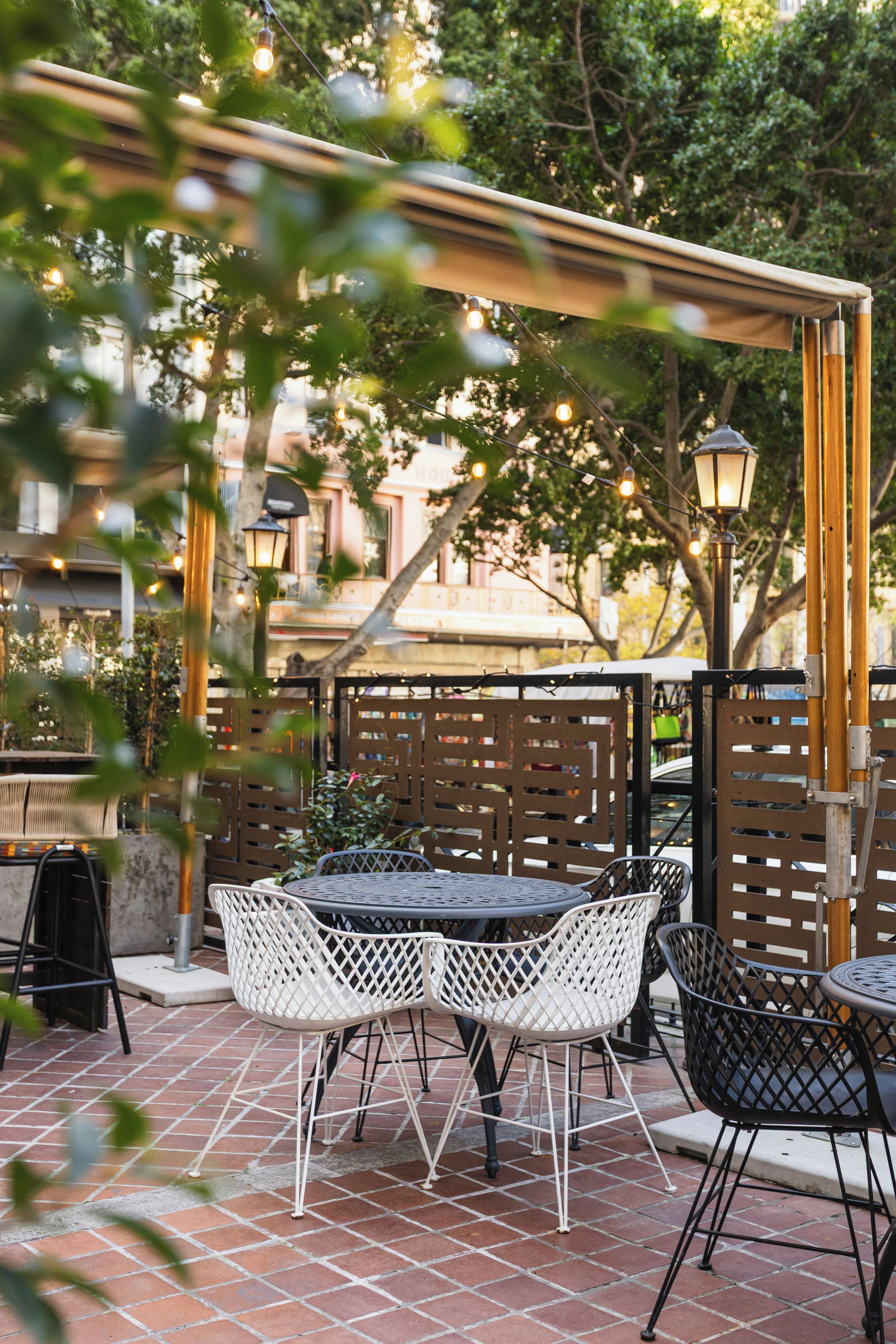 Un patio al aire libre con una mesa redonda y sillas blancas, rodeado por una pérgola de madera y luces colgantes.