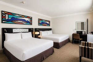 A hotel room with two beds, a desk, a chair, and two abstract paintings on the wall.