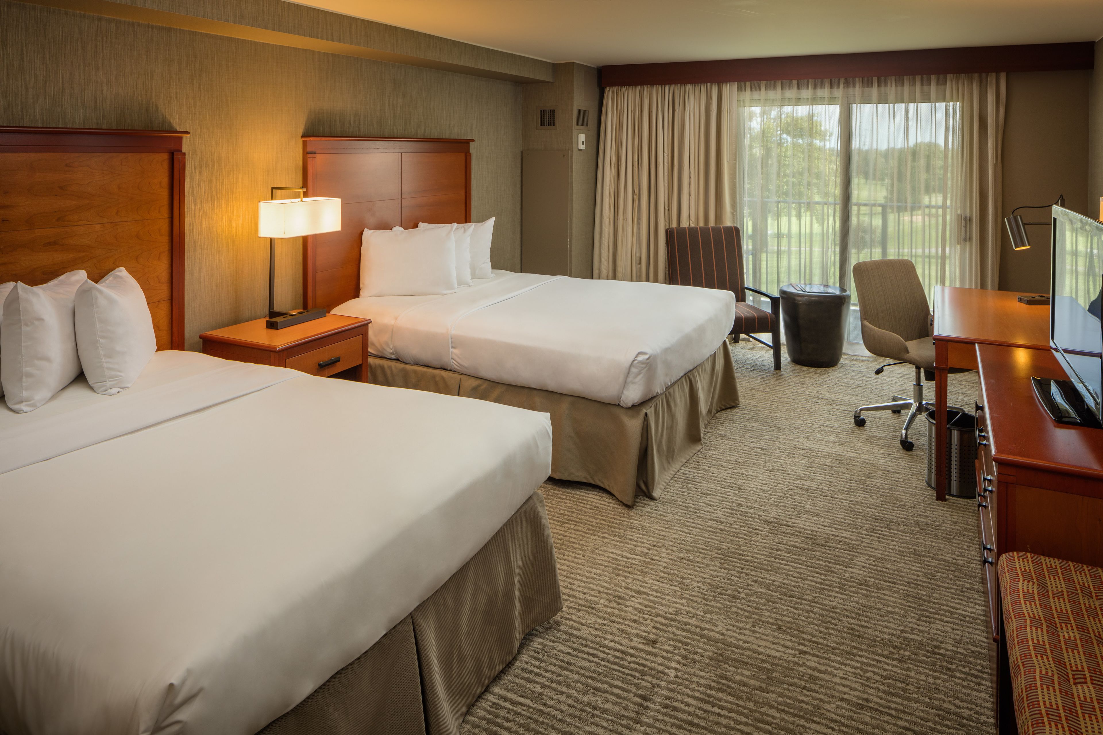 A hotel room with two beds, a desk, a chair, a lamp, and a large window with curtains.