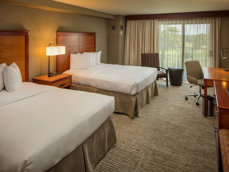 A hotel room with two beds, a desk, a chair, a lamp, and a large window with curtains.