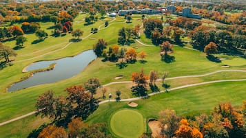 An aerial view of a golf course with a pond, surrounded by autumn-colored trees and buildings in the background.