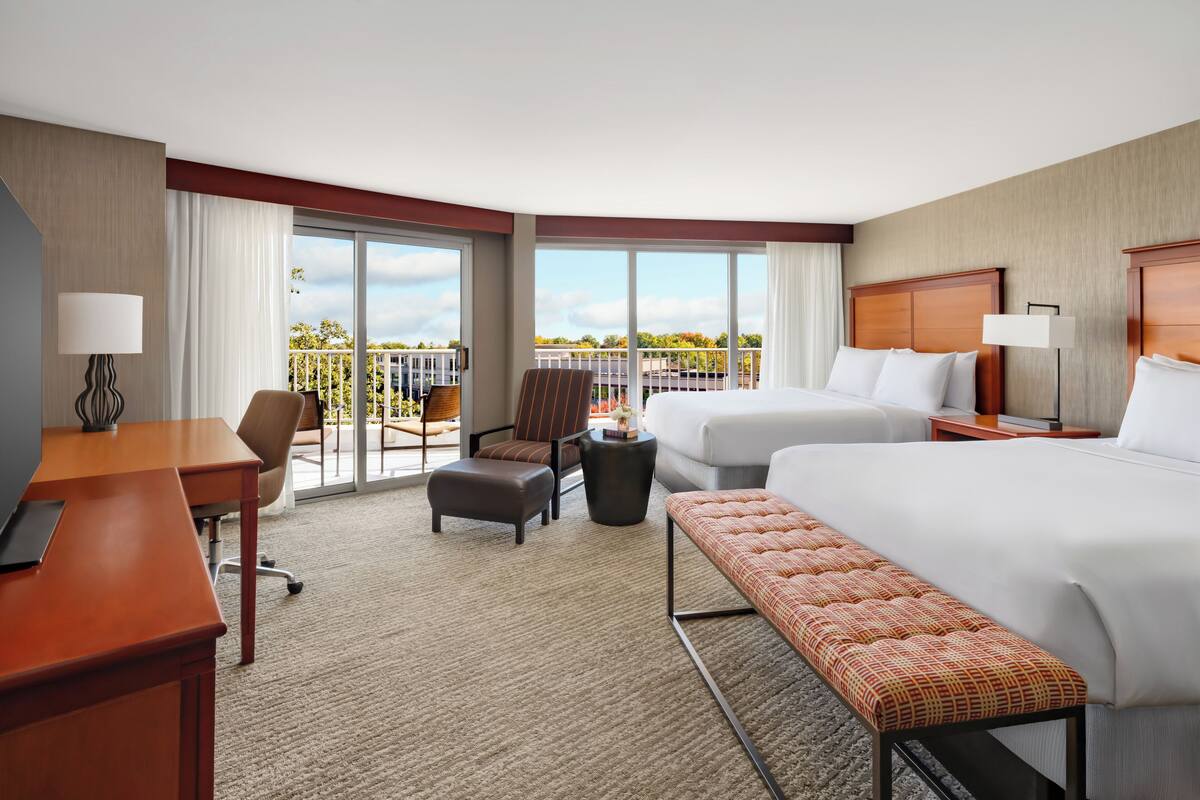 A hotel room with a large bed, a desk, a chair, a bench, and a balcony with a view of the city.