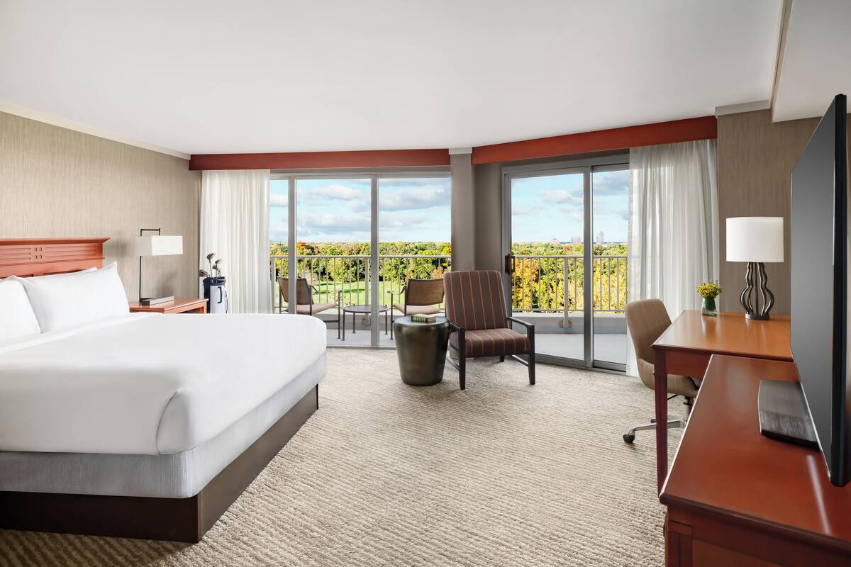 A hotel room with a large bed, a desk, a chair, and a balcony with a view of greenery.