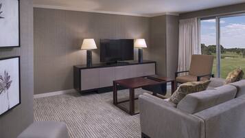 Habitación de hotel con televisor de pantalla plana sobre un mueble, dos lámparas de pie, un sofá, una silla y un amplio ventanal con vistas a un campo de golf.