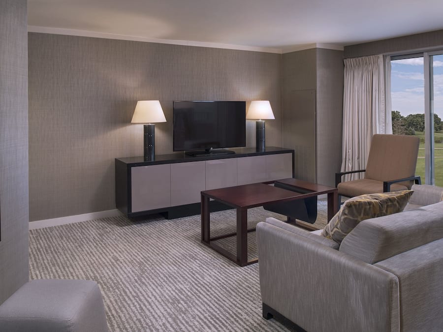 A hotel room with a flat-screen TV on a cabinet, two floor lamps, a sofa, a chair, and a large window with a view of a golf course.