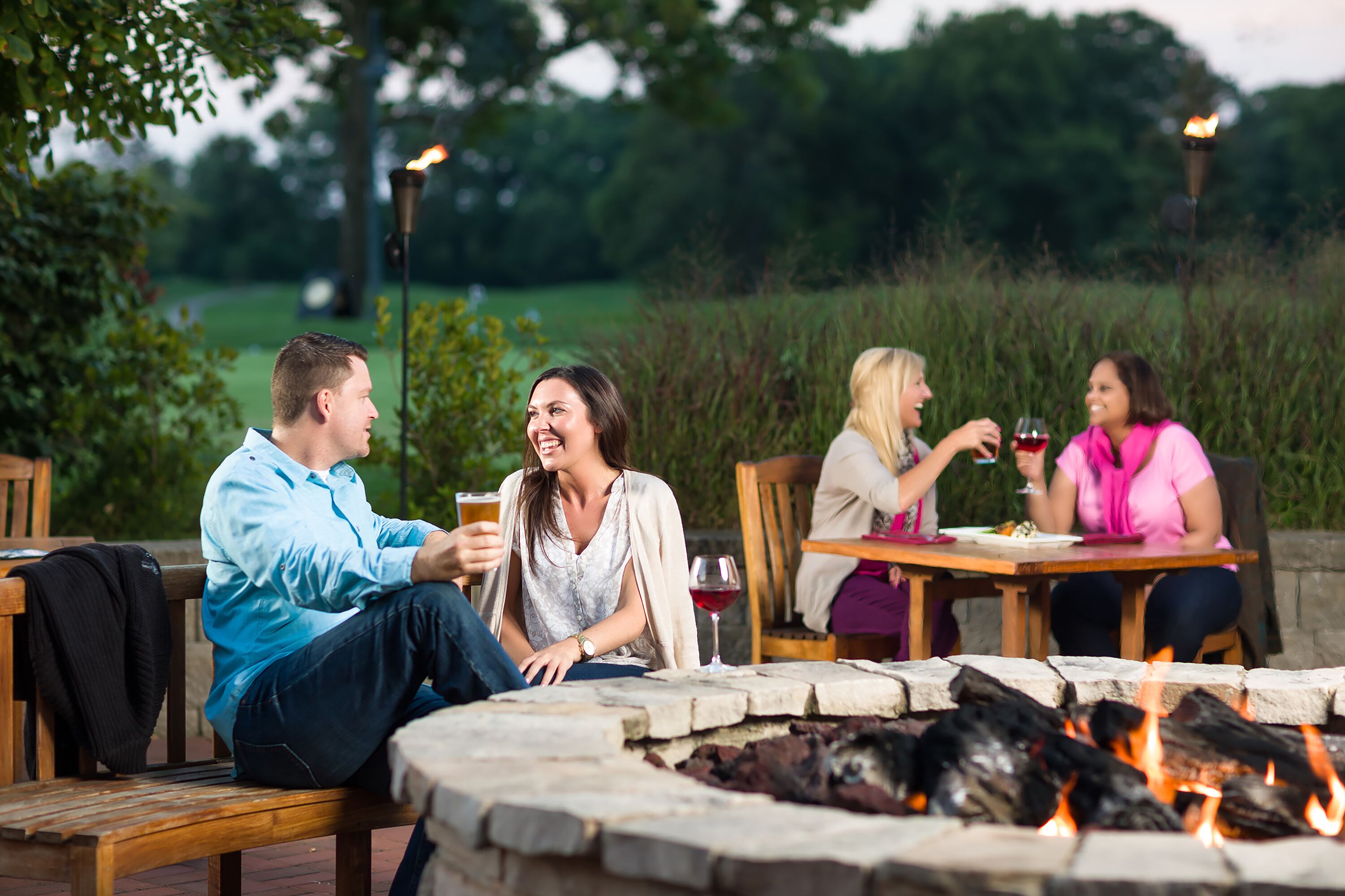 A group of people sitting around a fire pit with a grassy outdoor area in the background.
