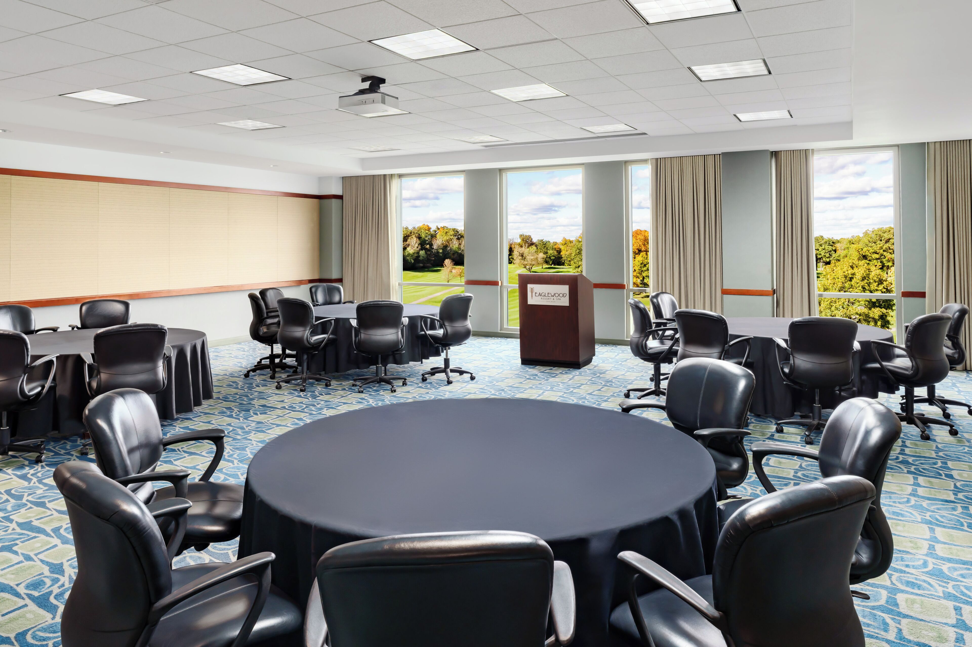 A conference room with a large round table, multiple chairs, a podium, and large windows with a view of trees.