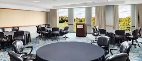 A conference room with a large round table, multiple chairs, a podium, and large windows with a view of trees.