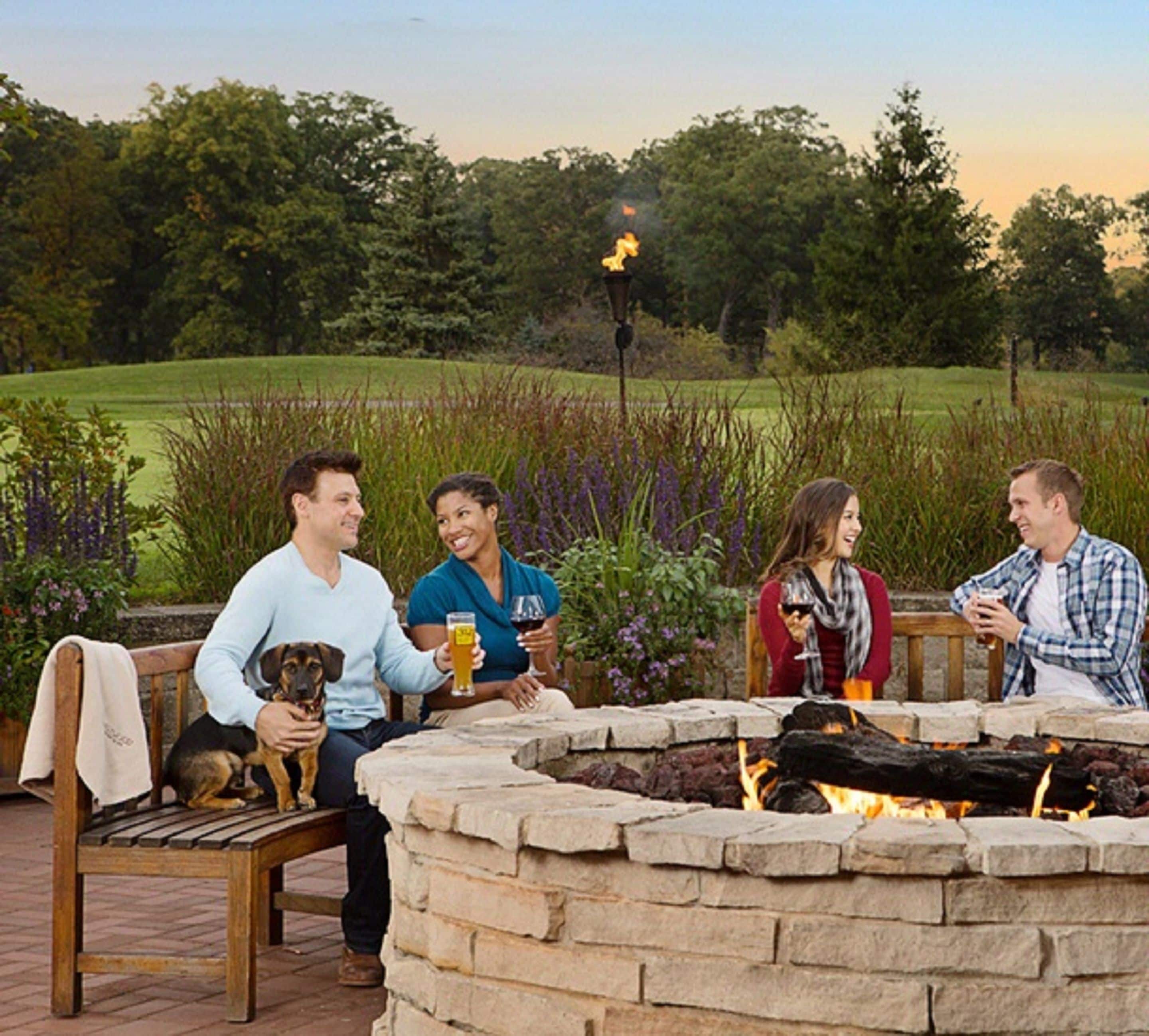 A group of people seated around a stone fire pit on a patio with a grassy field in the background.