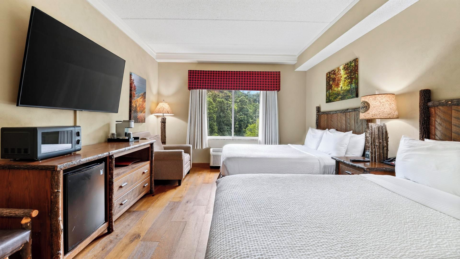 A hotel room with a flat-screen TV, a desk with a microwave, two beds, a window with a view of trees, and a red checkered valance.