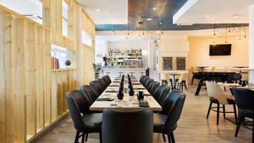 Un restaurant avec une longue table dressée pour un repas, un lambris en bois sur un des murs et un bar à l’arrière-plan.