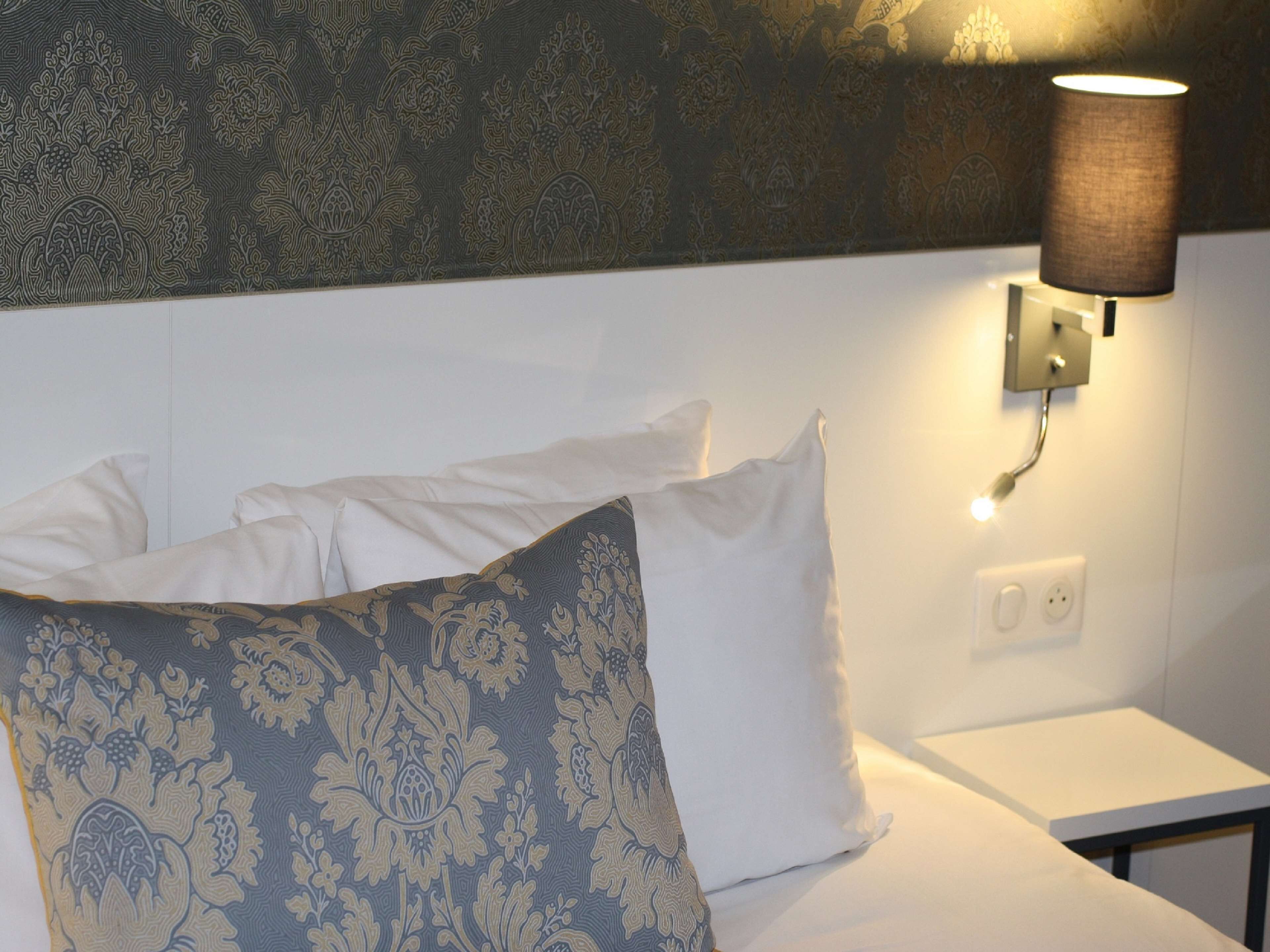 Een netjes opgemaakt bed met witte en gestreepte kussens, een wandlamp en een klein nachtkastje.