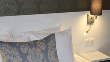 Een netjes opgemaakt bed met witte en gestreepte kussens, een wandlamp en een klein nachtkastje.