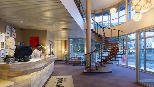 Un vestíbulo de hotel con una recepción, una escalera y amplias ventanas que ofrecen vistas al exterior.