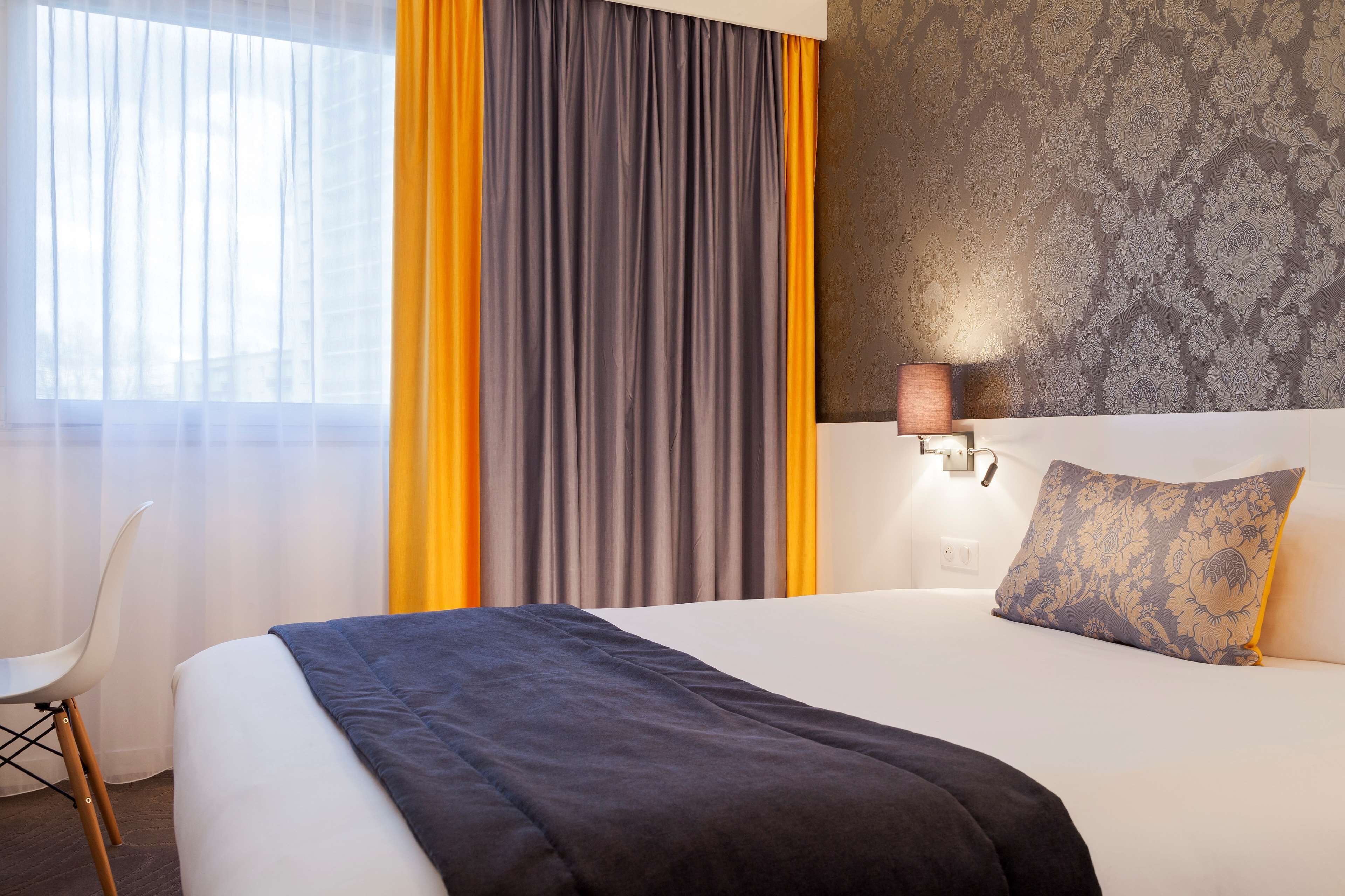 Quarto de hotel com cama, poltrona, cortinas, parede com papel de parede estampado e luminária.