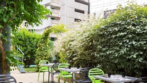 Un café al aire libre con mesas y sillas, rodeado de follaje y un edificio al fondo.