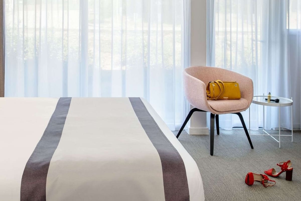 Habitación de hotel con una cama grande, una silla y un bolso amarillo, además de zapatos rojos en el suelo.