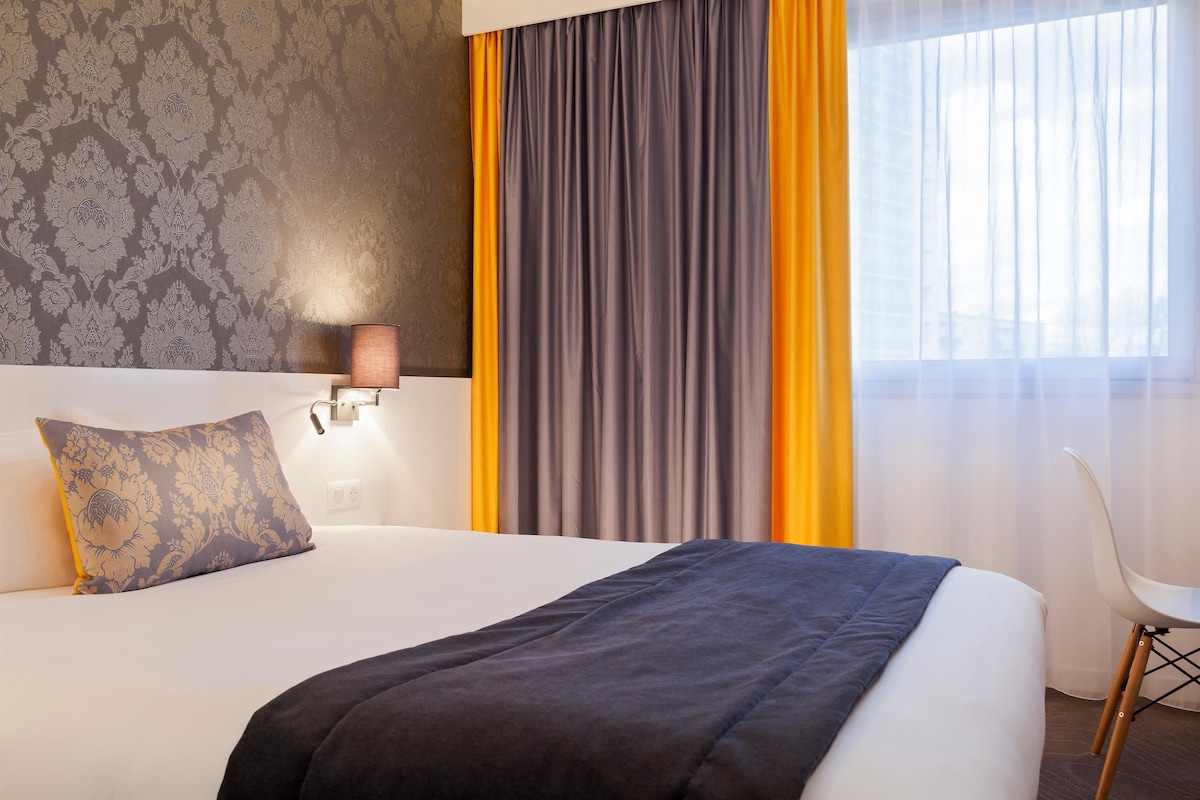 Habitación de hotel con cama, mesita de noche, lámpara, sillón y ventana con cortinas.