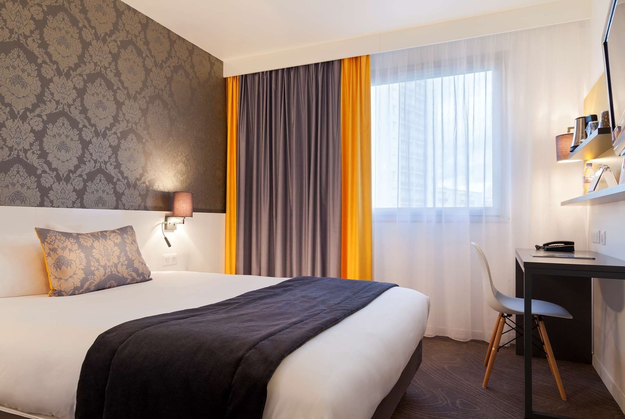 A hotel room with a bed, a desk, a chair, a lamp, and a window with curtains.