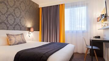 A hotel room with a bed, a desk, a chair, a lamp, and a window with curtains.
