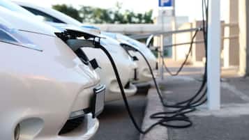 Varios vehículos eléctricos cargando en una estación, con un cartel de estacionamiento al fondo.