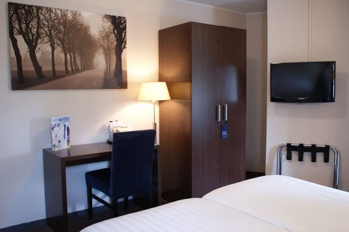A hotel room with a bed, a desk with a chair, a wardrobe, a television mounted on the wall, and a lamp.