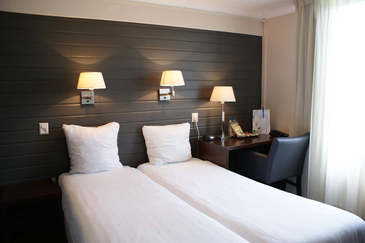 A hotel room with two beds, a desk with a chair, and two wall-mounted lamps.