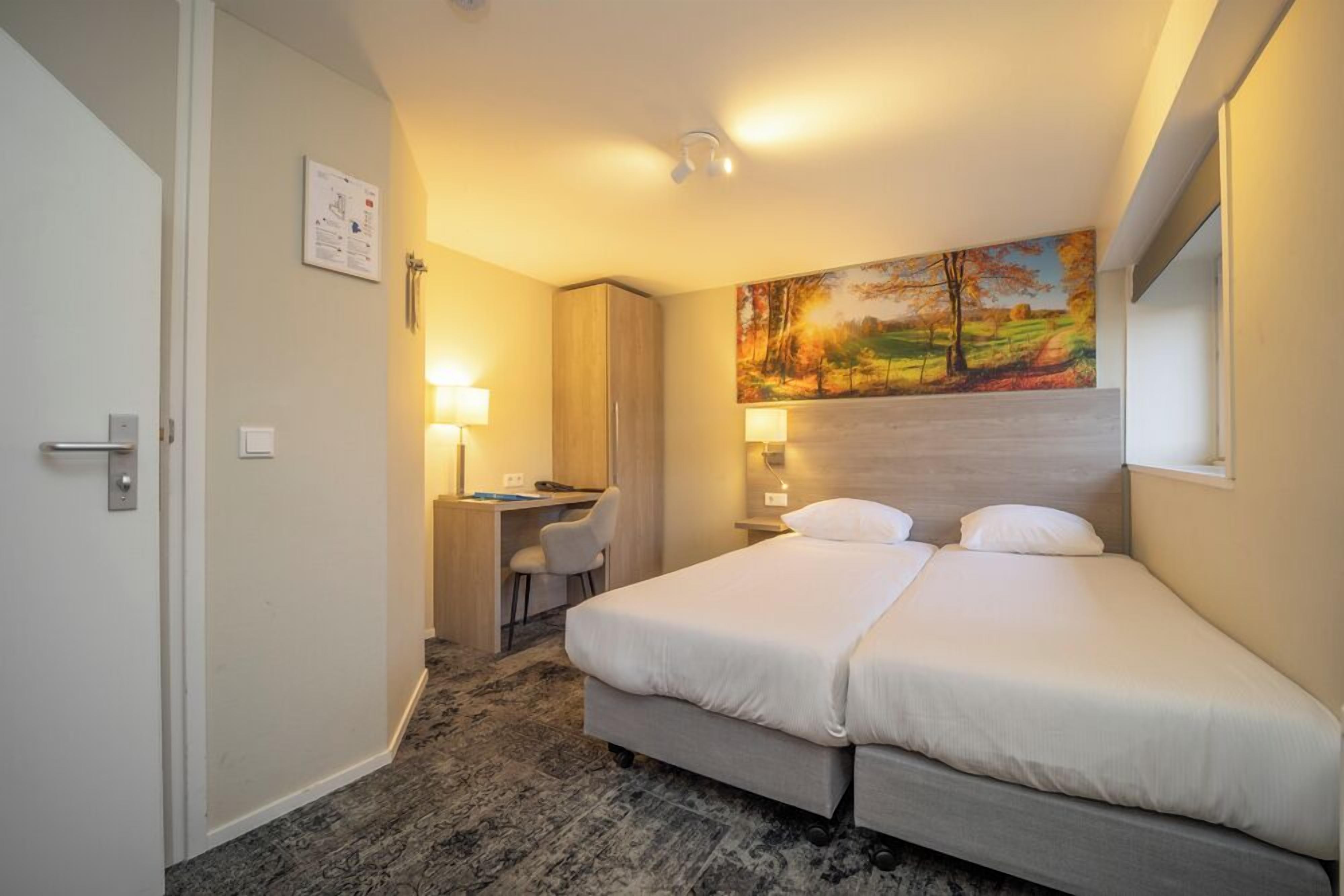 A hotel room with a bed, desk, chair, and a painting on the wall.