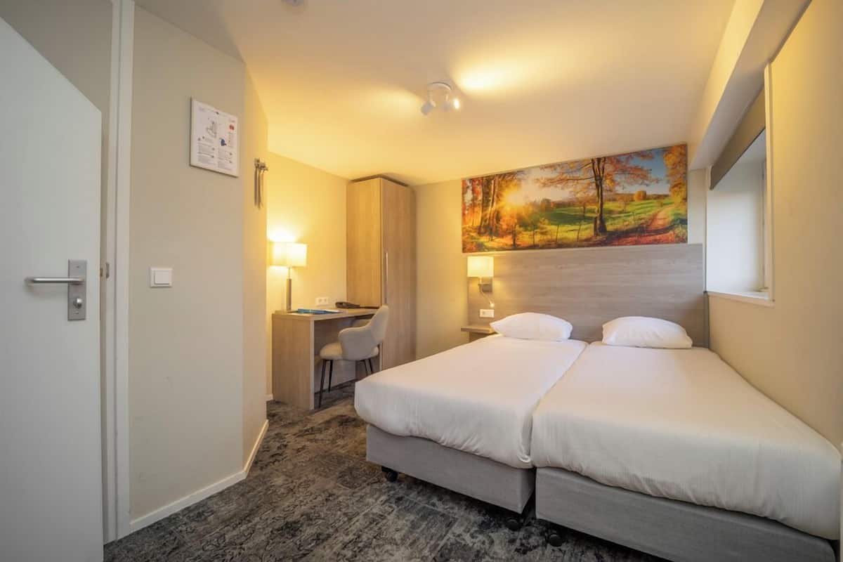 A hotel room with a bed, desk, chair, and a painting on the wall.