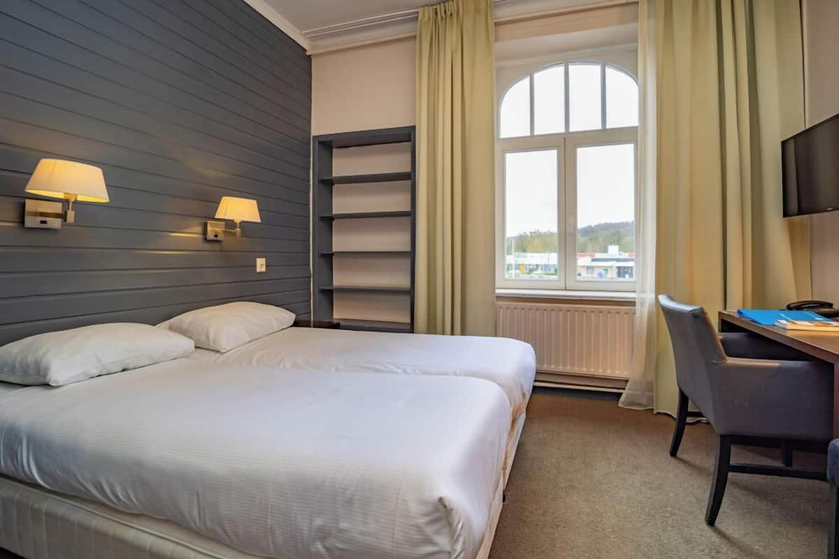 A hotel room with a bed, two bedside lamps, a desk with a chair, a TV, and a window with curtains.