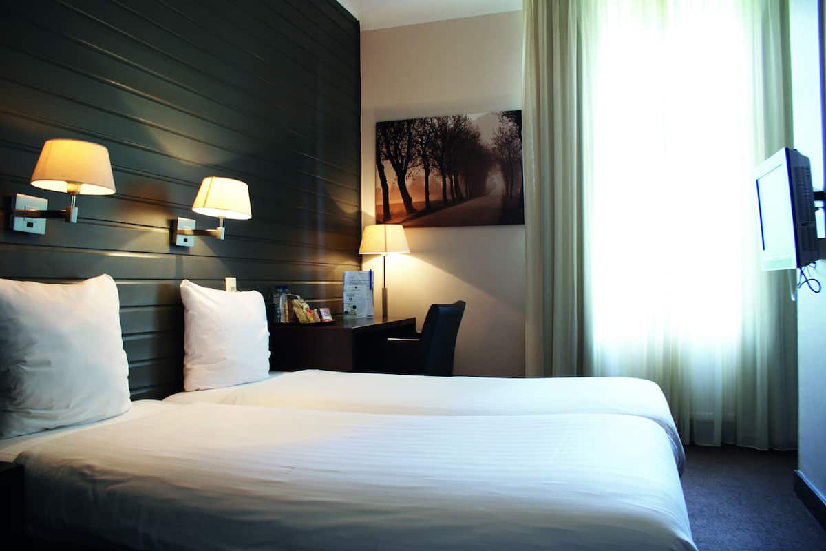 A hotel room with a large bed, two wall-mounted lamps, a desk with a chair, a television, and a painting on the wall.