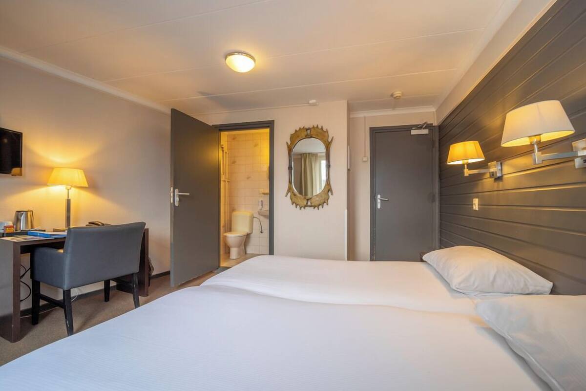 A hotel room with a bed, a desk, a chair, a bathroom, and a mirror.