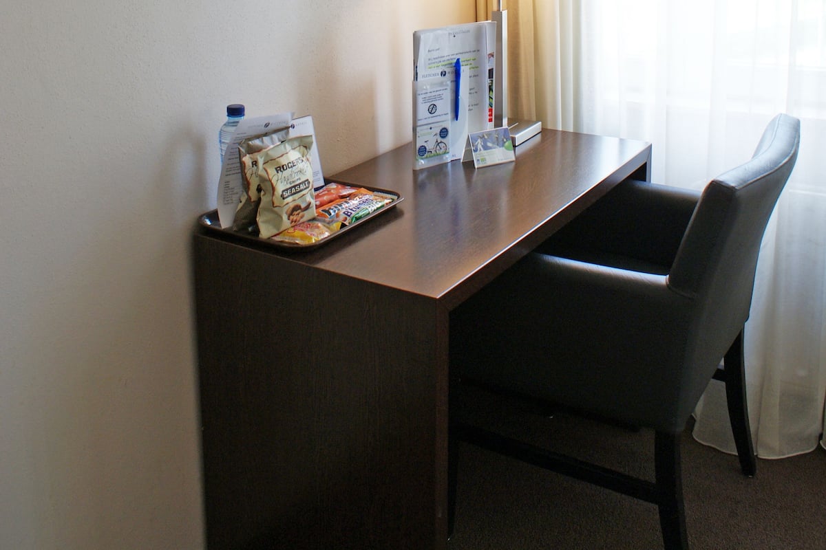 A hotel room with a desk, chair, lamp, and a painting of trees.